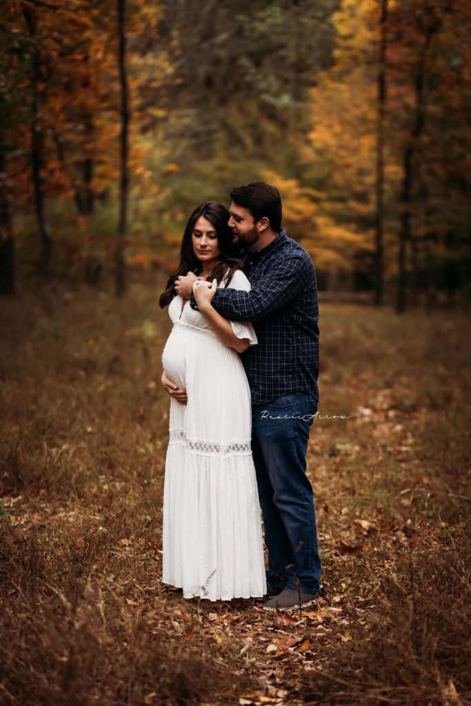 Husband grasping onto his wife and whispering into her ear during their maternity session surrounded by fall foliage.