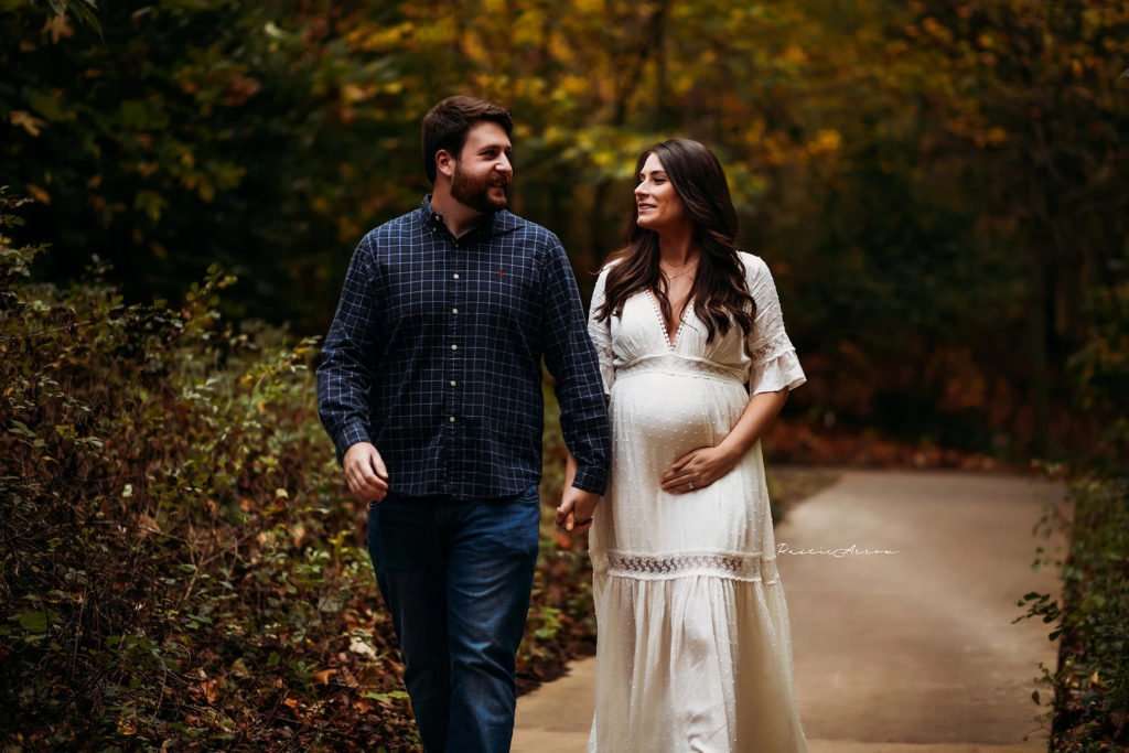 Husband and wife walking down trail surrounded by fall leaves, while wife is holding her pregnant belly