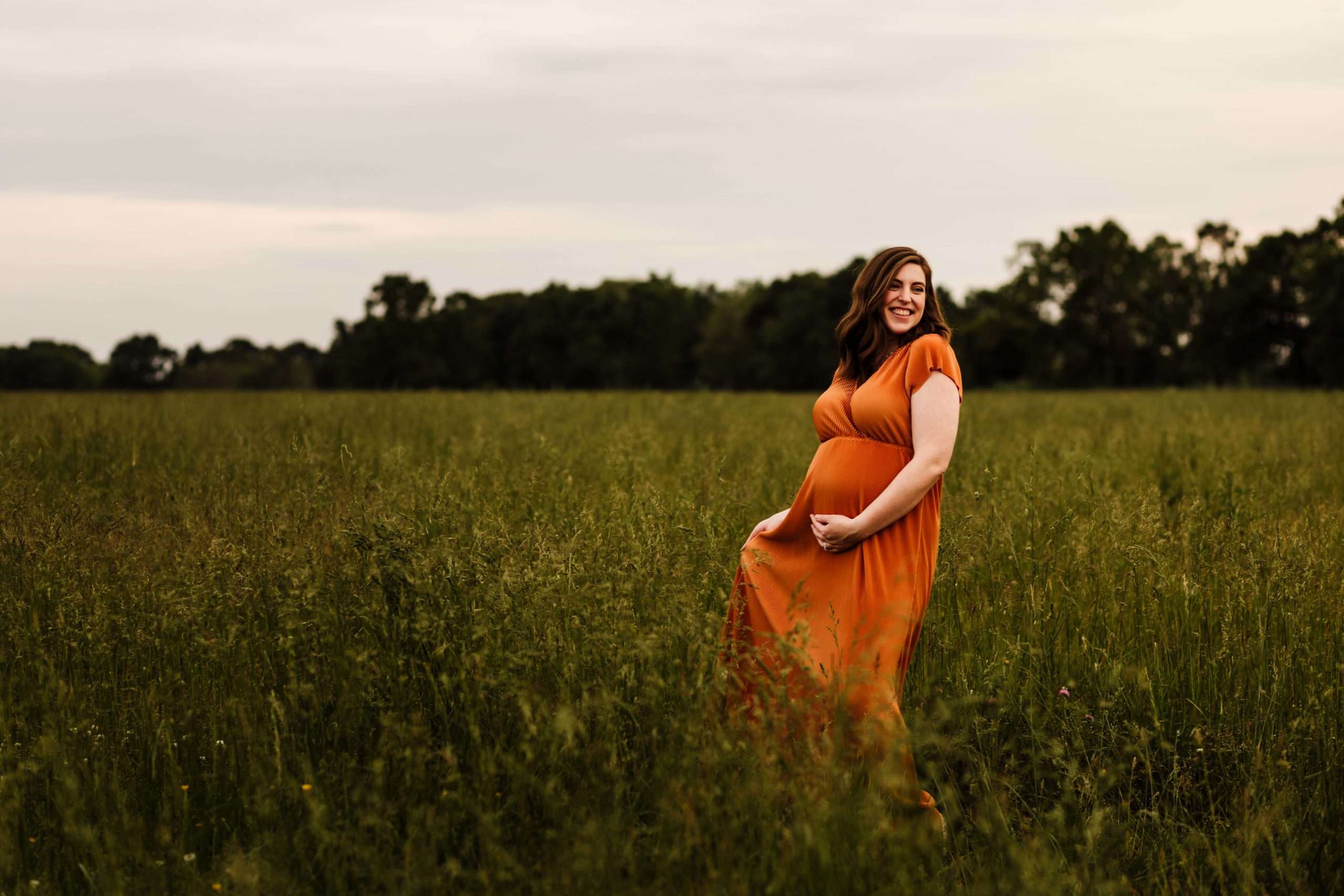 Pregnant woman standing in a tall grass field in a rust colored dress.
