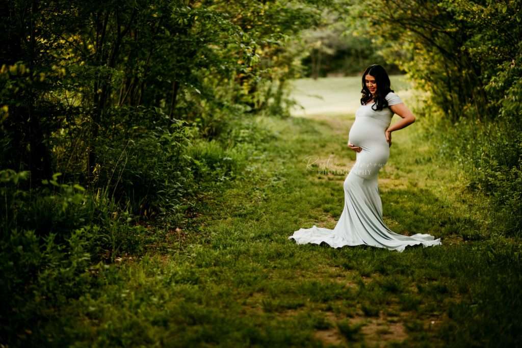 Pregnant woman cradling her belly under trees on a trail in Cave Springs