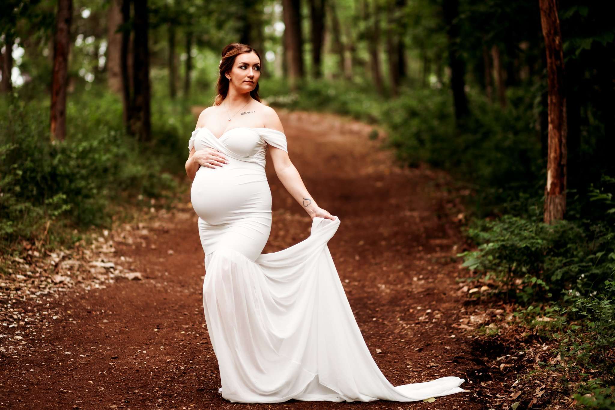 pregnant woman in white gown in woods and holding train of gown