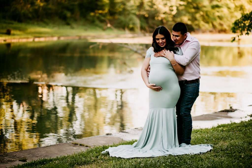 Pregnant couple expecting new baby girl while standing next to a beautiful watershed area. Mom is wearing a light green dress while cradling her belly and dad is holding her and looking down at belly