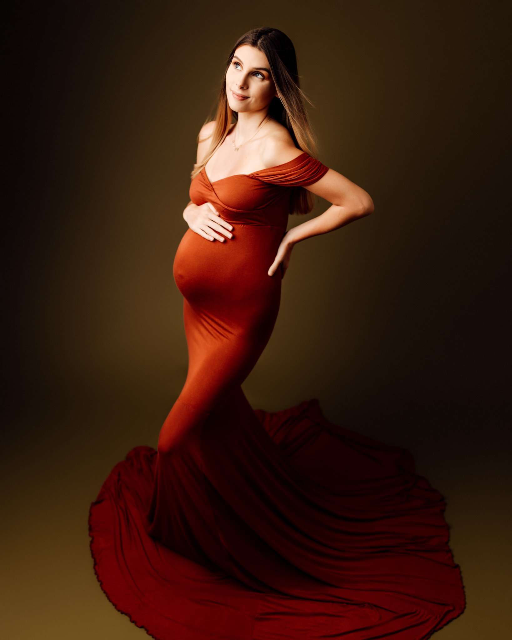 pregnant woman in studio wearing copper mermaid style dress with hand on baby bump