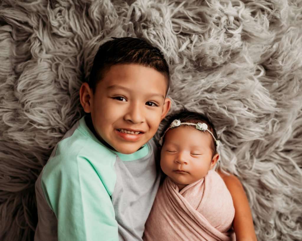 brother holding wrapped little sister on gray flokati rug during newborn photography session