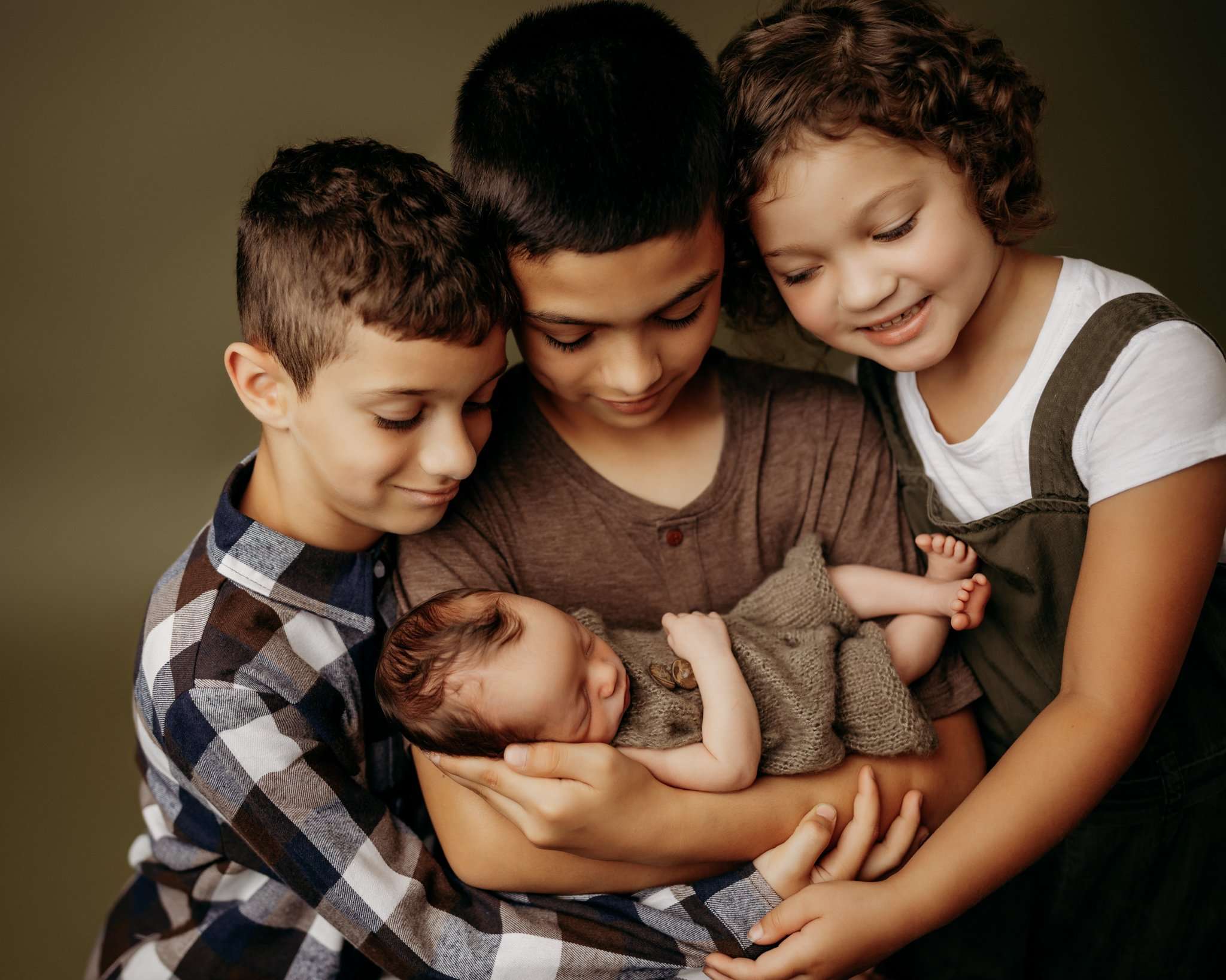 siblings holding baby brother on dark green backdrop while looking at their sibling