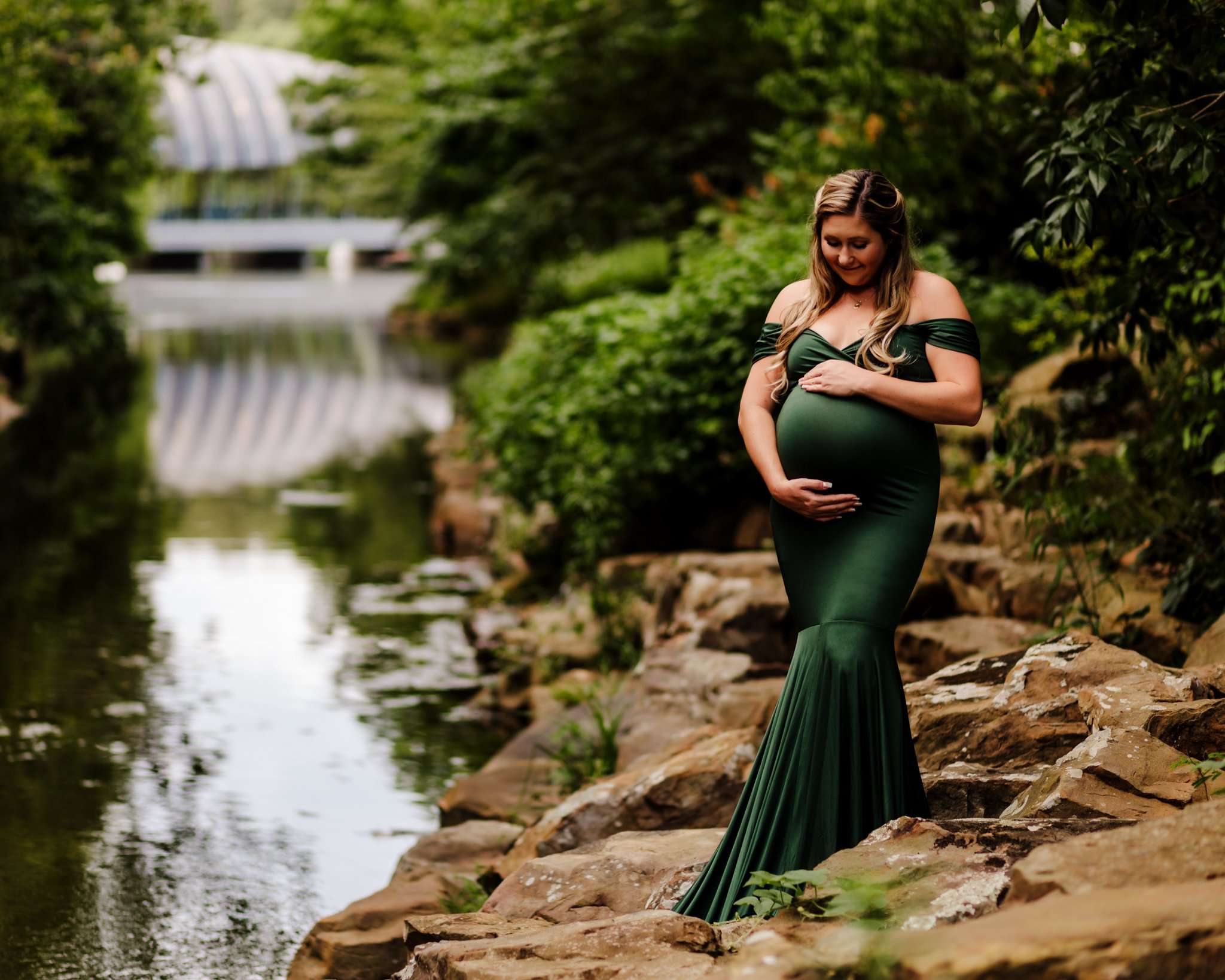 Pregnant woman standing on rocks beside a creek at Crystal Bridges Museum, cradling her belly and looking down at her baby with Crystal Bridges in the background.