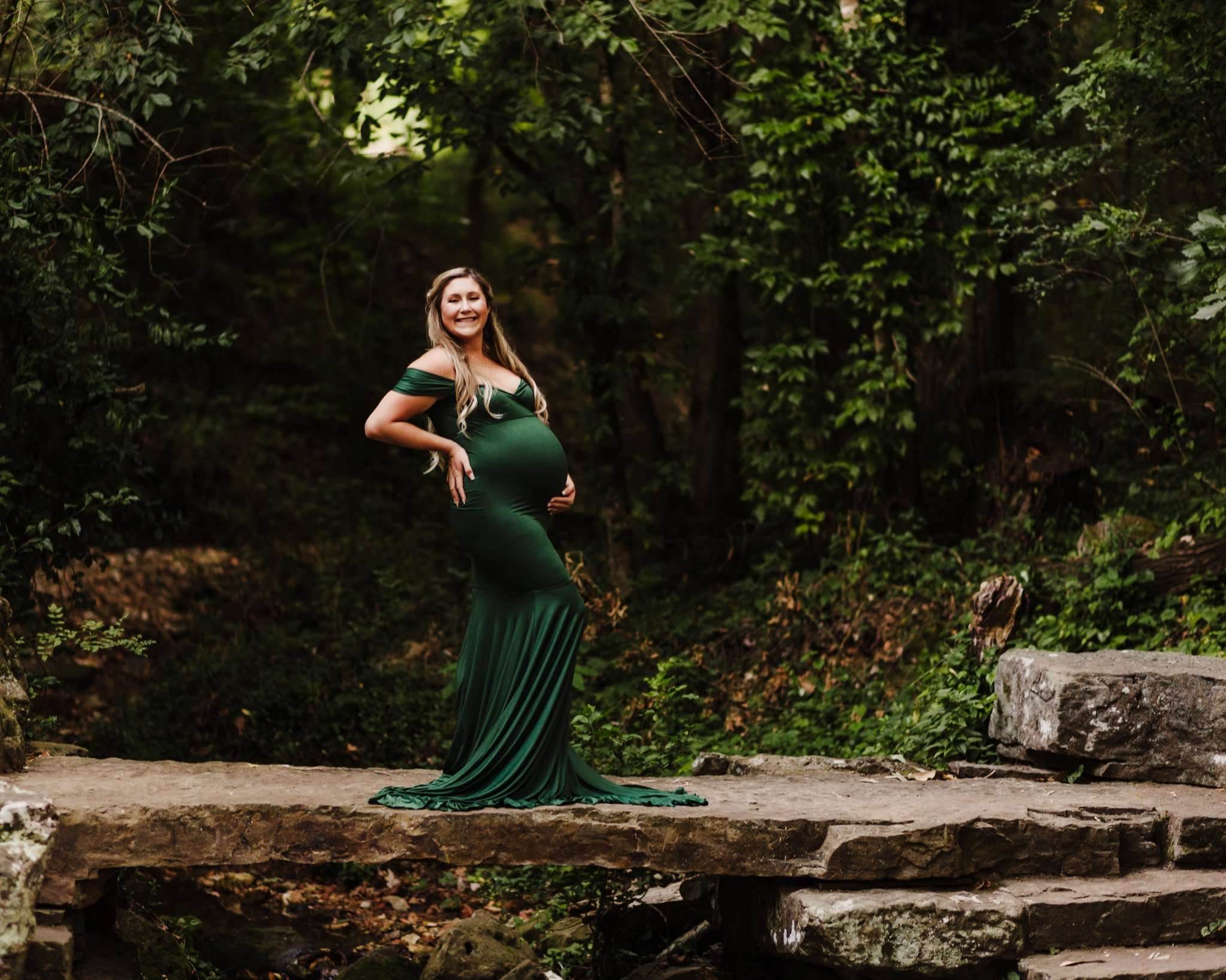 maternity photography session at Crystal Bridges along the art trail on rock ledge. Mom is holding belly and smiling at camera surrounded by foliage