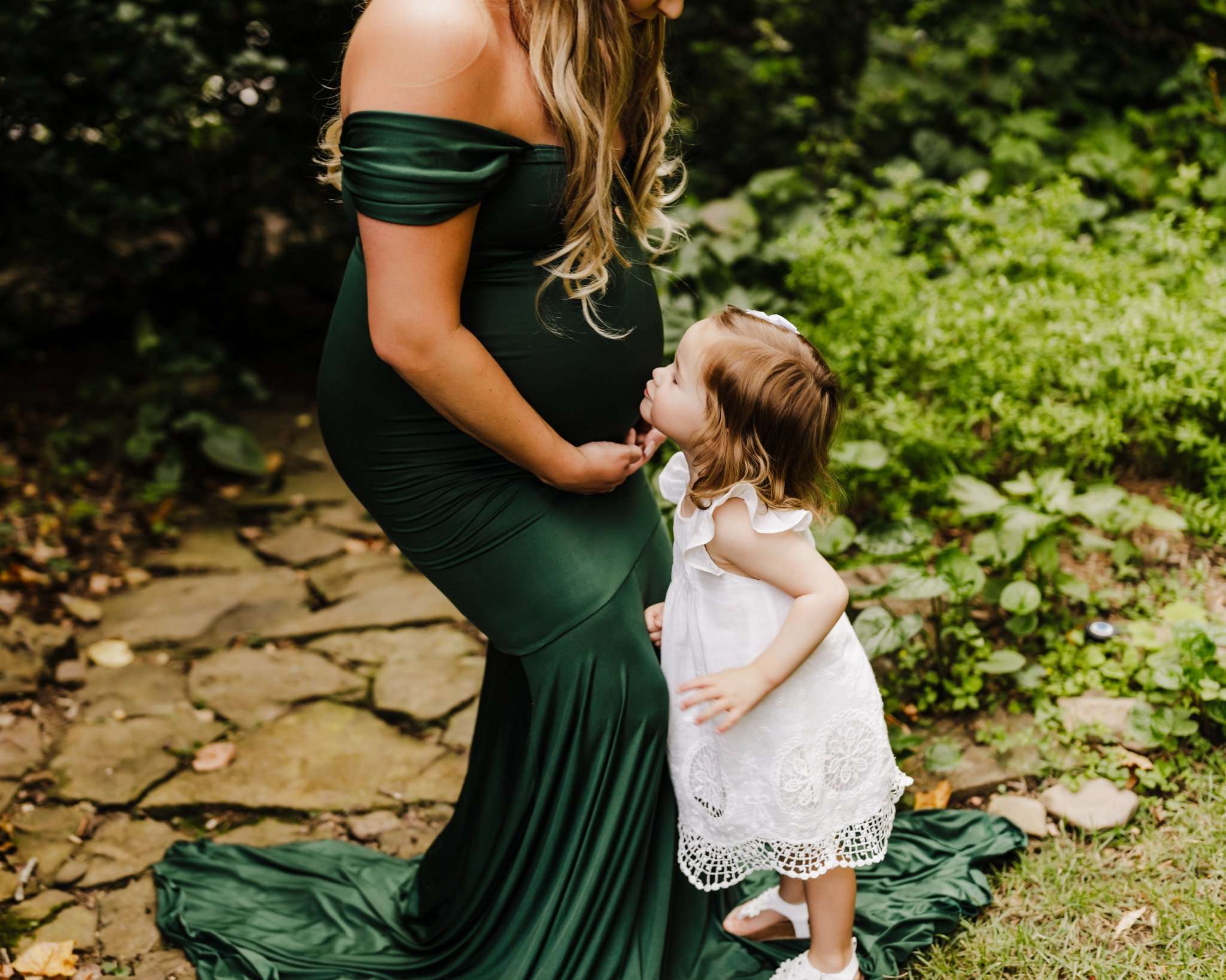pregnant woman in green dress leaning over for her daughter to kiss her belly in wooded area