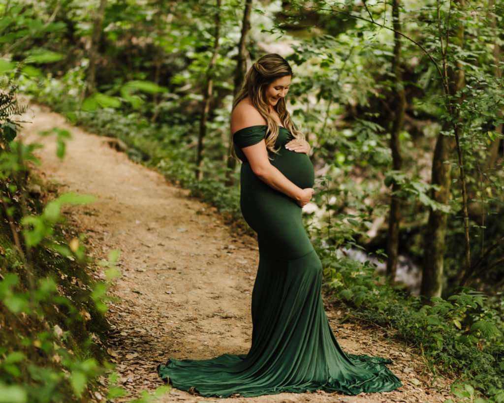 pregnant mom standing on trail surrounded by foliage while looking down and cradling her baby belly.