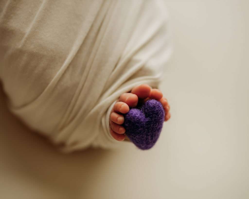 newborn photography session on white backdrop with purple heart being held by baby's feet