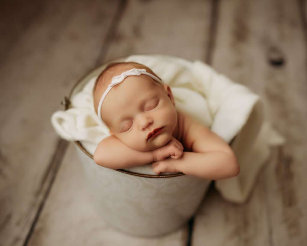 newborn in bucket with white cover on it, wearing a white bow, while sleeping