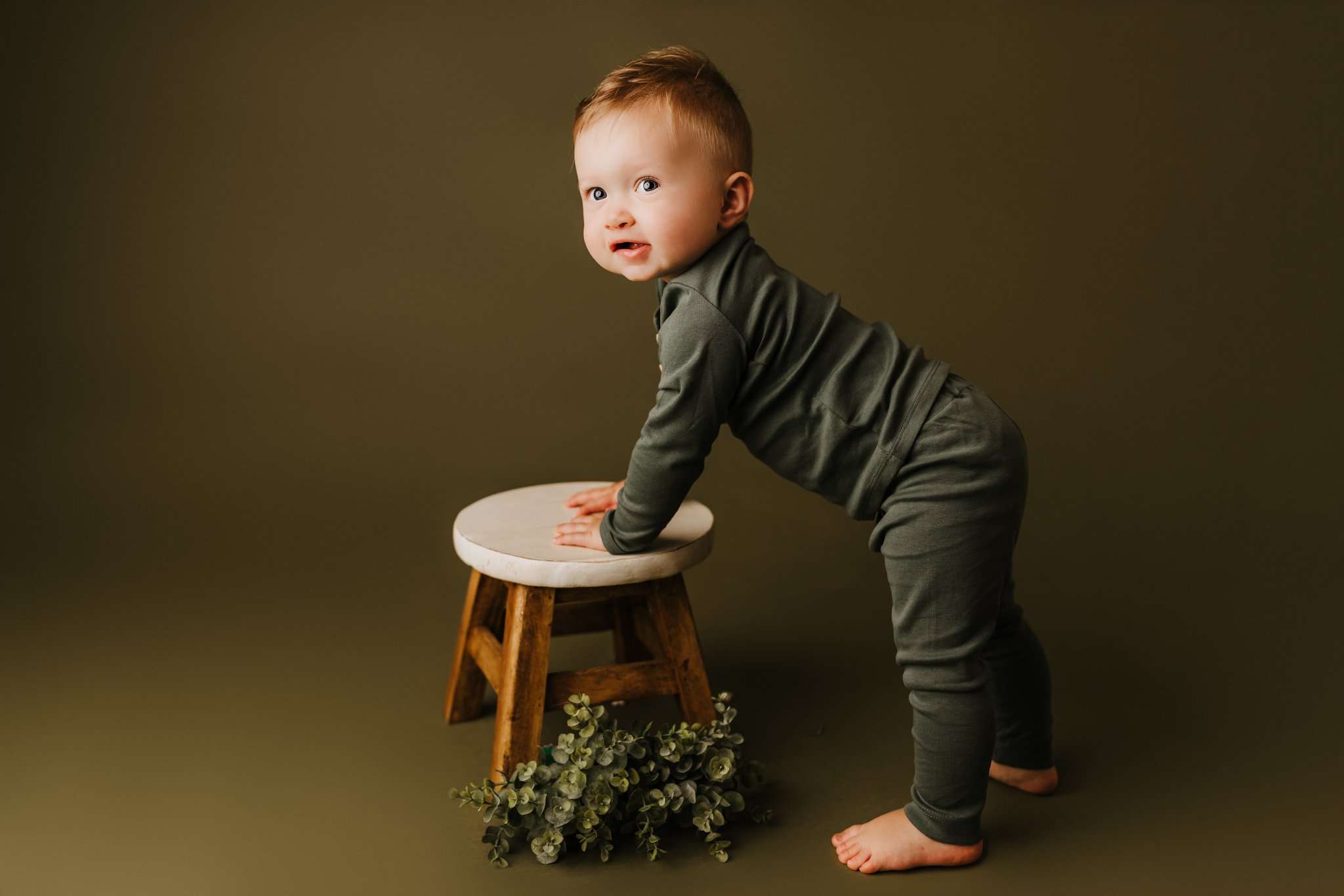 one year old leaning on stool while in front of green backdrop wearing green clothes and smiling at camera