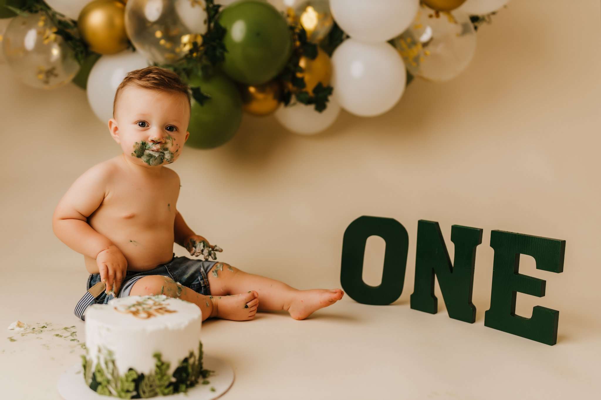 one year old enjoying his cake in studio on a cream backdrop in front of green, gold, and white balloon garland