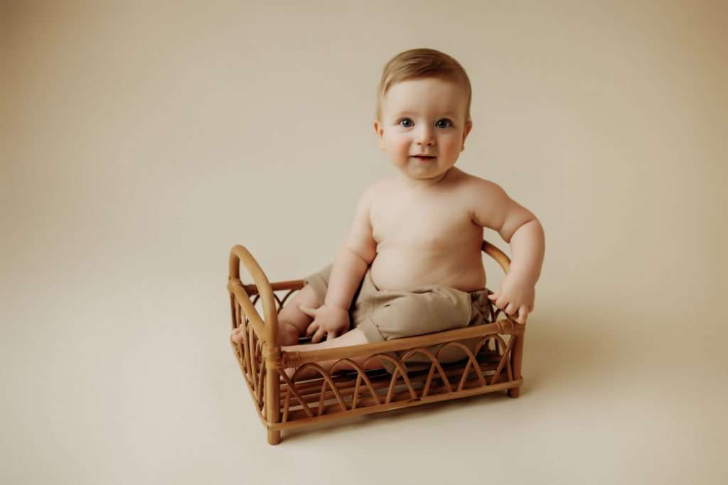 Infant in bamboo bed looking at the camera smiling on a cream colored backdrop