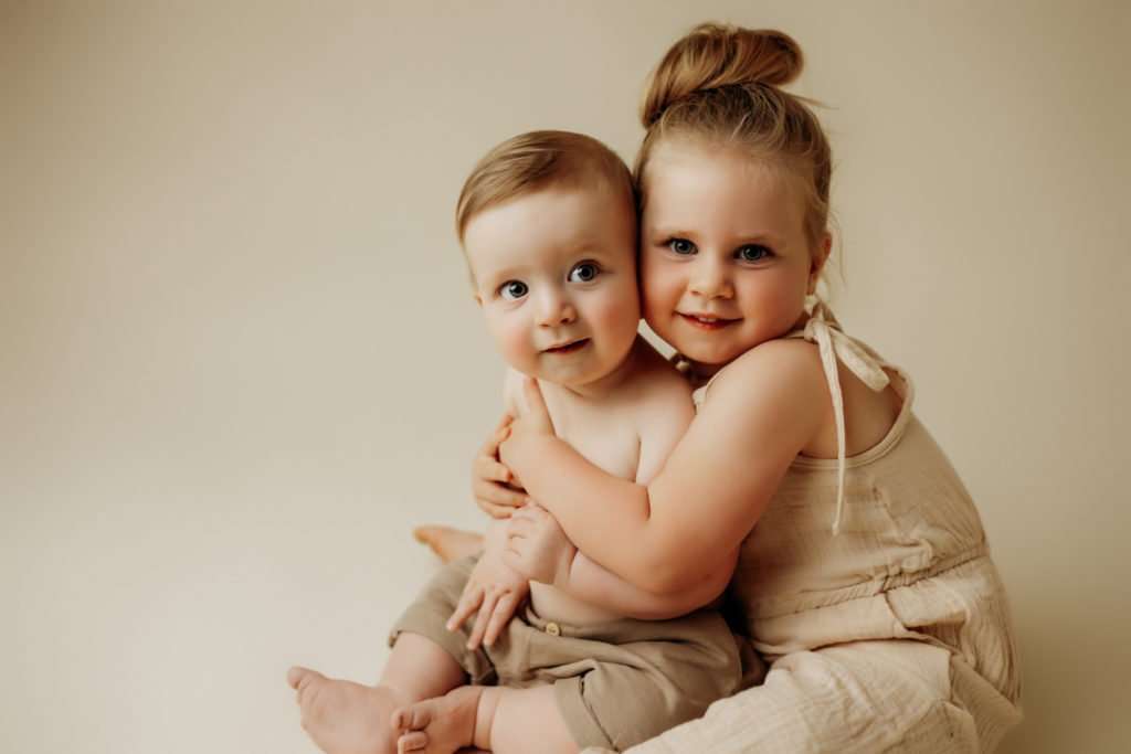 Brother and sister hugging on cream colored backdrop while smiling at camera
