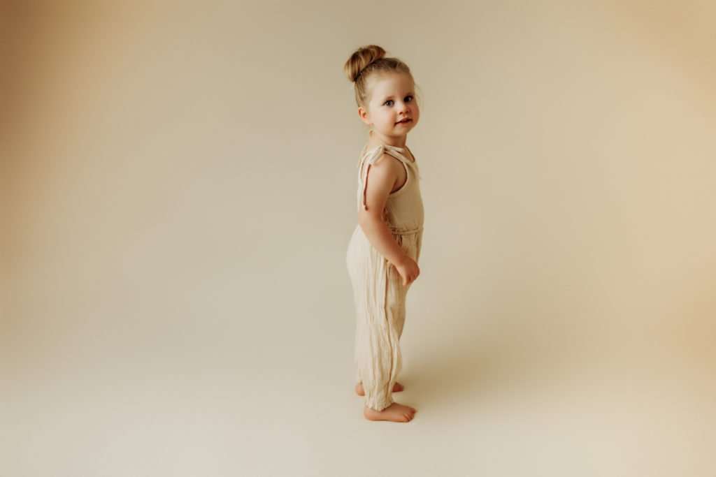 Toddler standing on cream colored backdrop with matching romper looking at camera