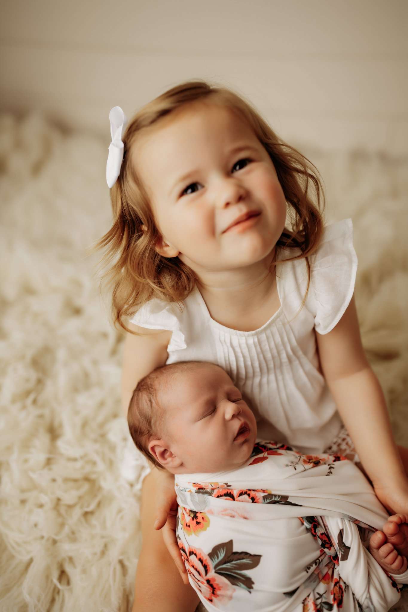 nwa newborn wrapped in a white and floral wrap sleeping while in big sisters arms while big sister looks and smiles at the camera