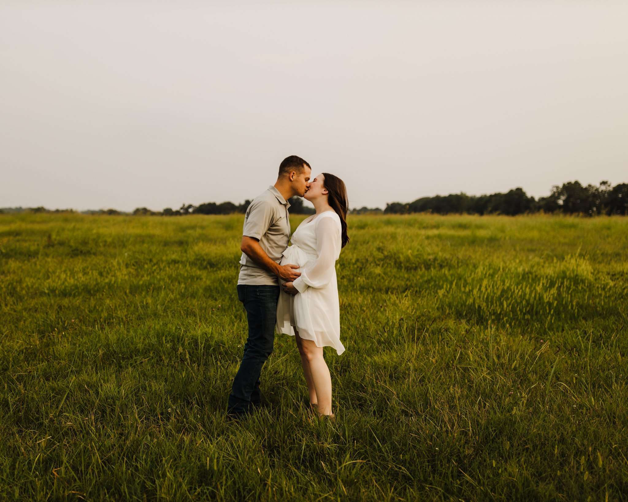maternity photography session with mom kissing dad and mom in a white dress in an open field