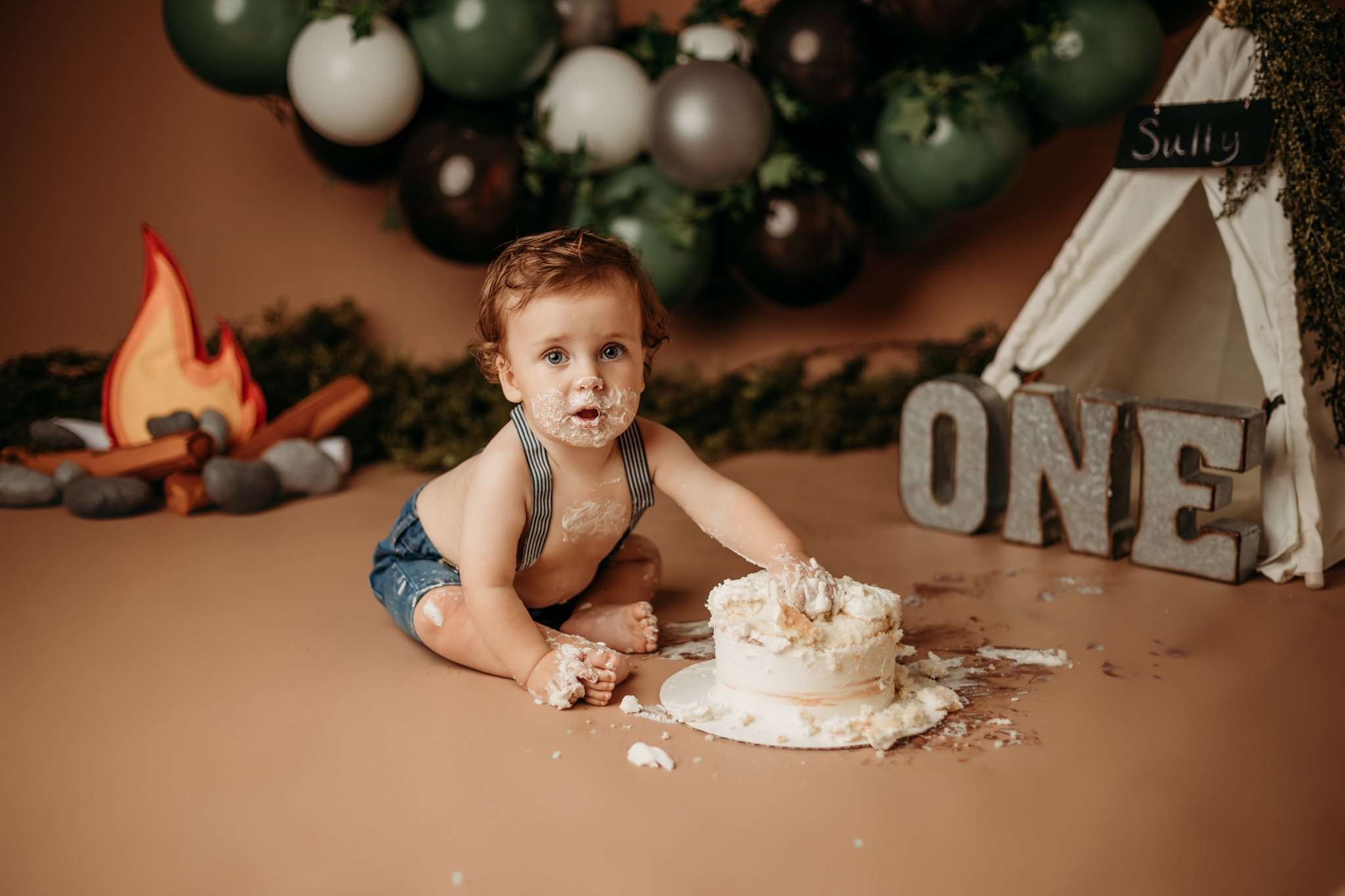 NWA one year images in studio with toddler on brown backdrop with a tent behind him and him eating cake while looking at the camera