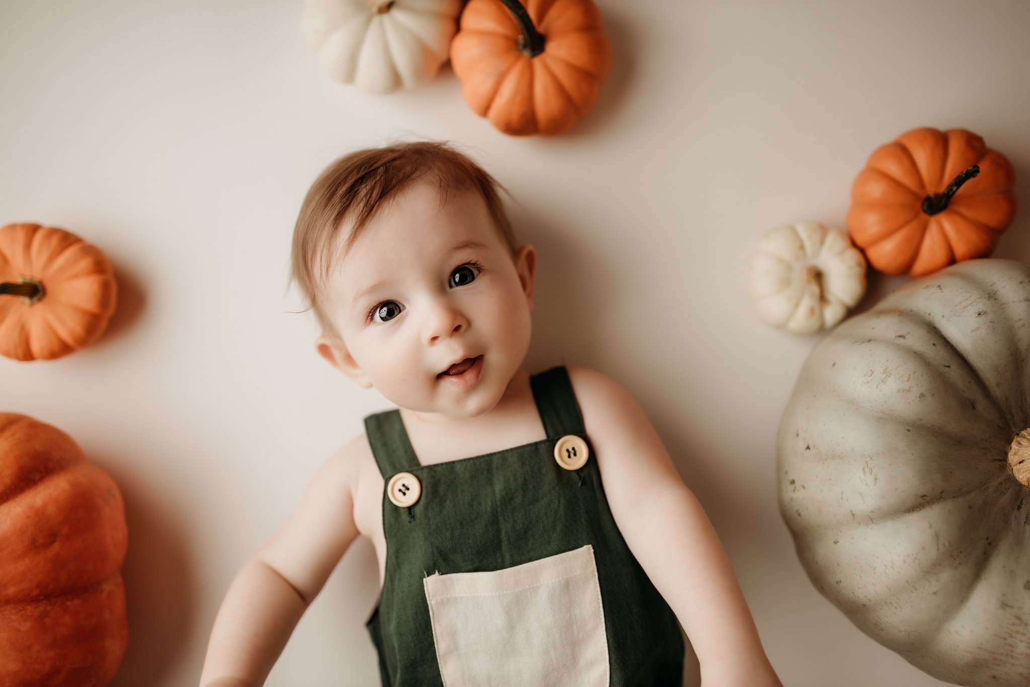 6 month old child laying on white background and surrounded by pumpkins while wearing green overalls