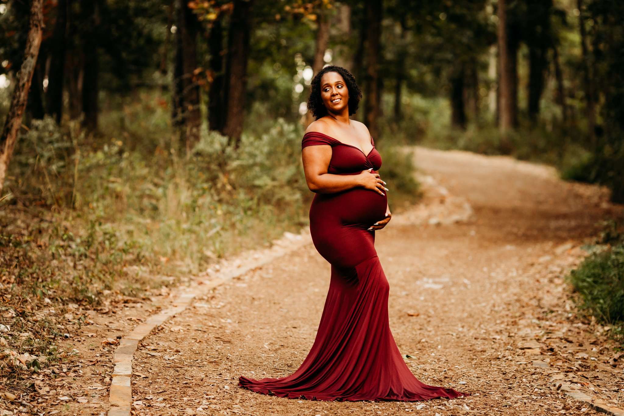 pregnant woman standing on trail surrounded by trees in a red gown cradling her baby belly and smiling