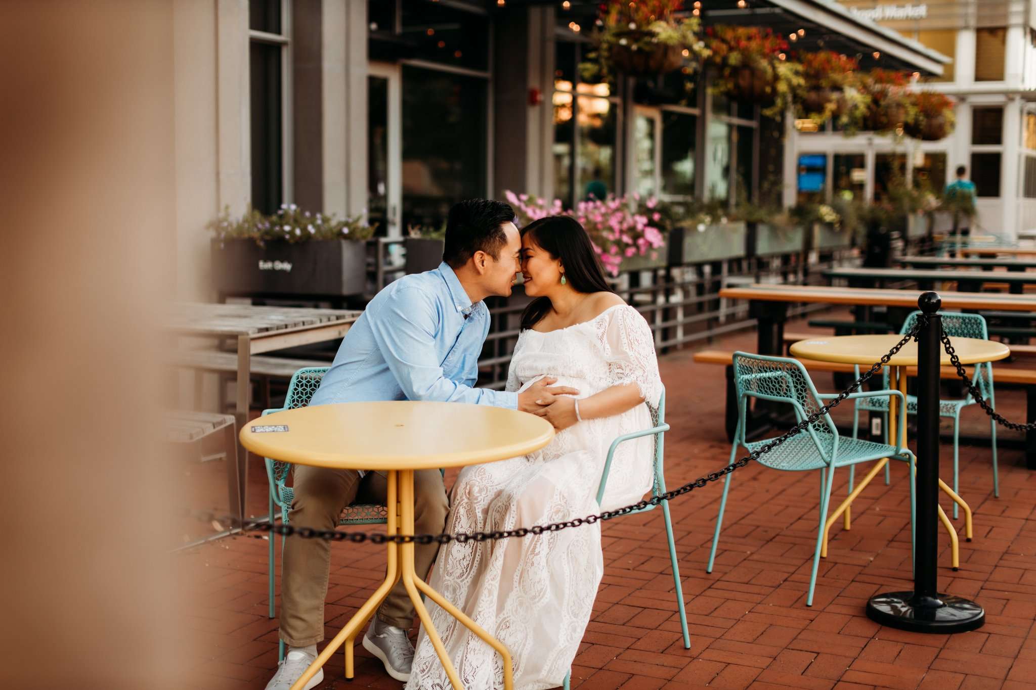husband and wife sitting at a yellow streetside table going in for a kiss while holding their baby belly.