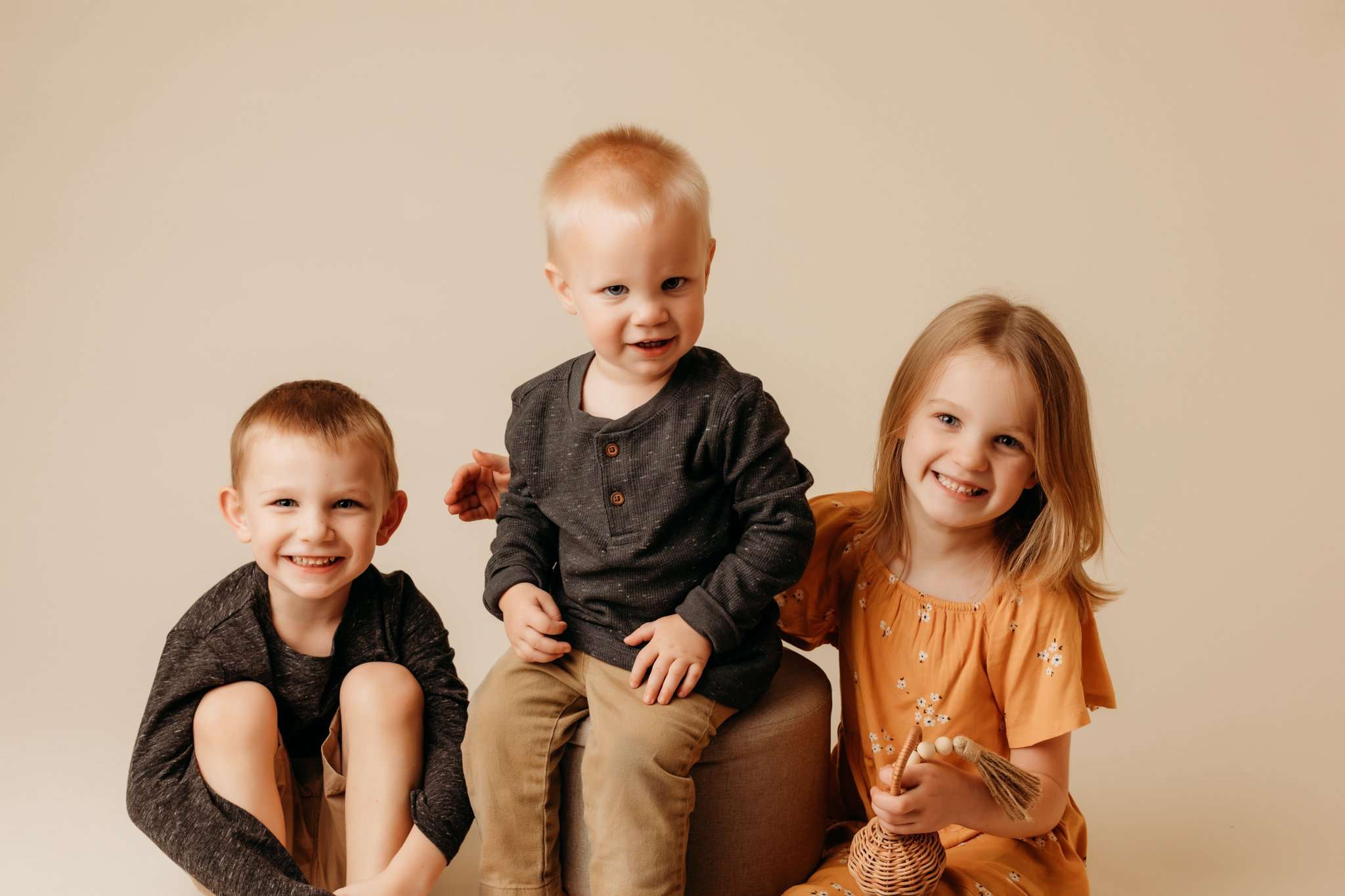 three children sitting on a cream backdrop smiling at the camera