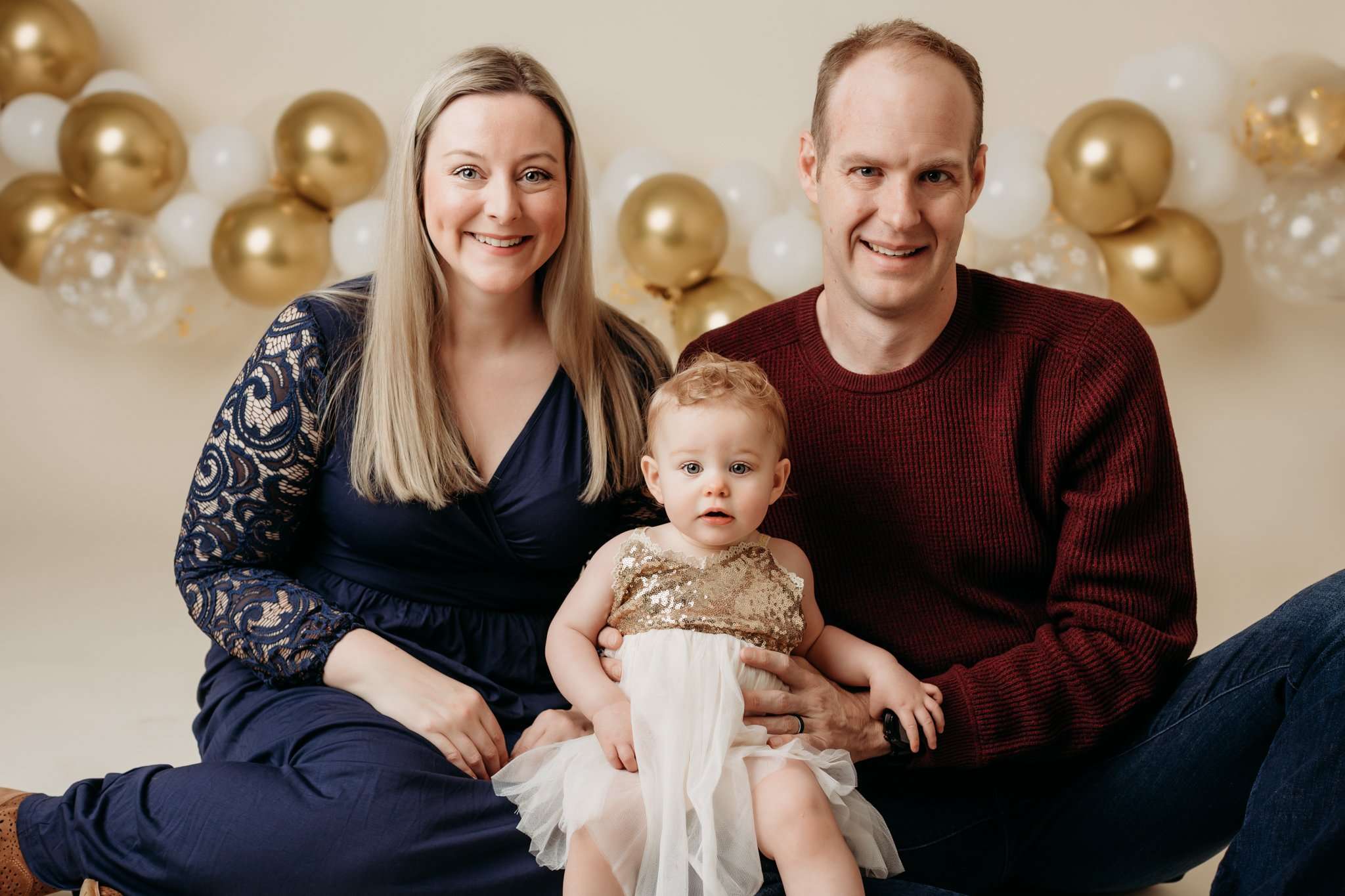 parents and child looking at the camera and smiling on a cream backdrop with balloons in the background