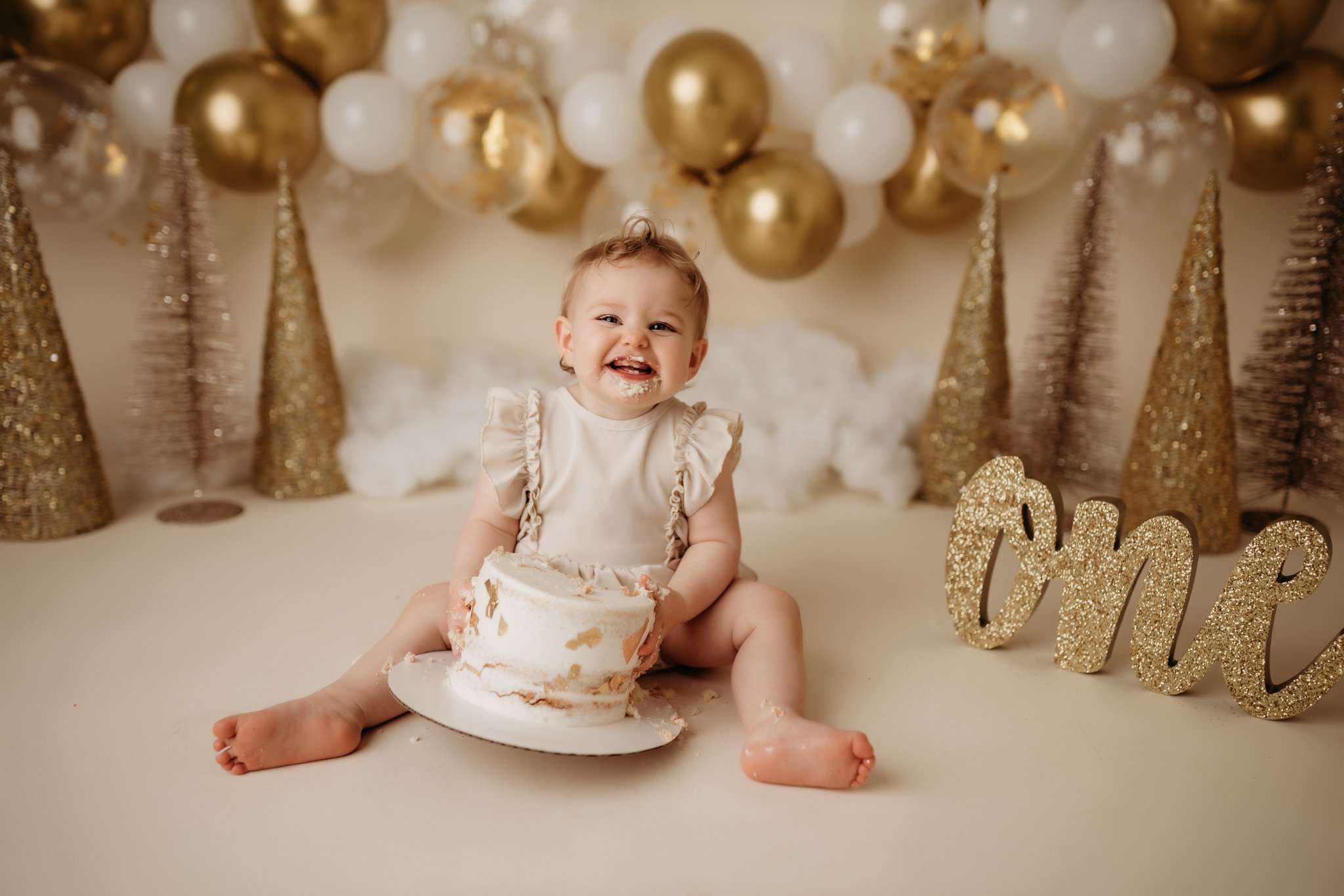 one year milestone photography session with girl on a cream backdrop with her smiling at the camera in a cream romper while eating her cake with trees and balloon garland in the background