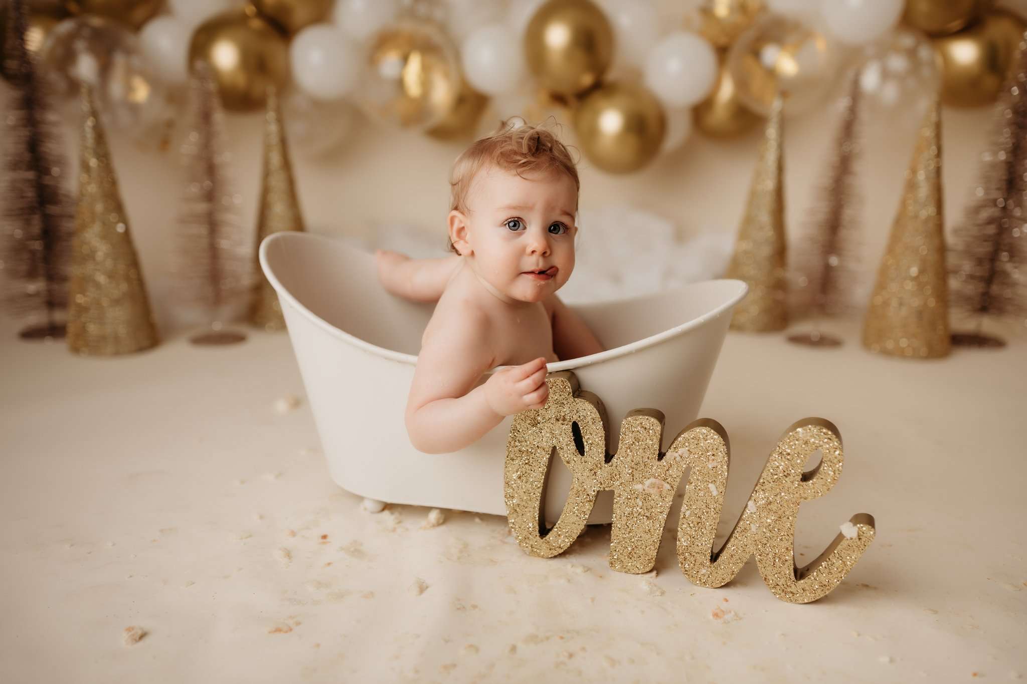 baby girl in bathtub next to a gold glitter one sign on a cream backdrop with a gold and white balloon garland in the background