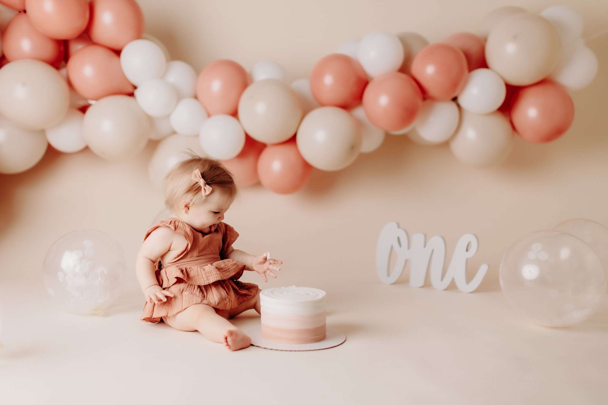 one year old girl on a cream backdrop with pink and cream balloons behind her, poking at a cake in front of her.