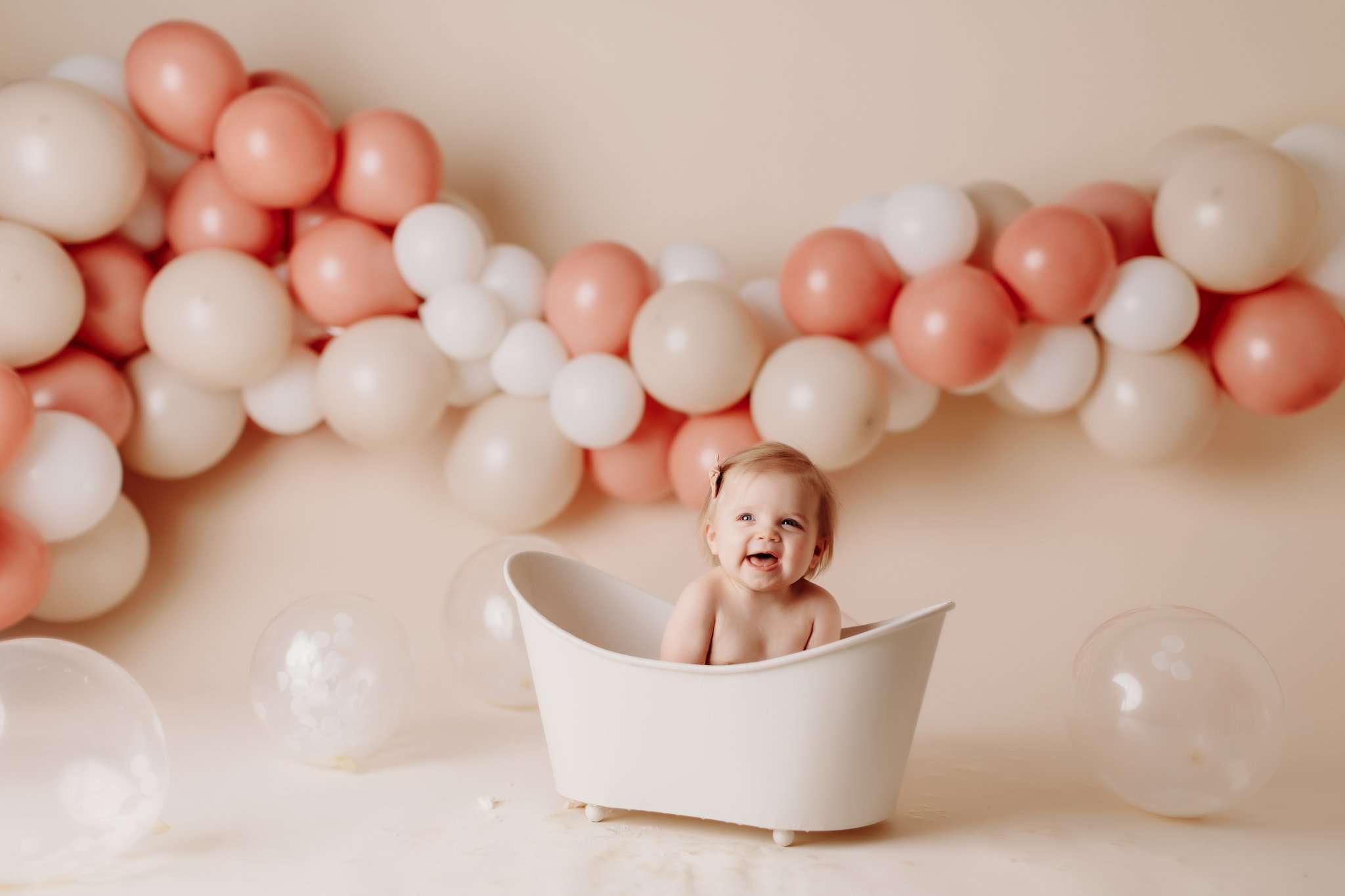 One year old in a tub on a cream backdrop surrounded by pink and cream balloons smiling and laughing at the camera