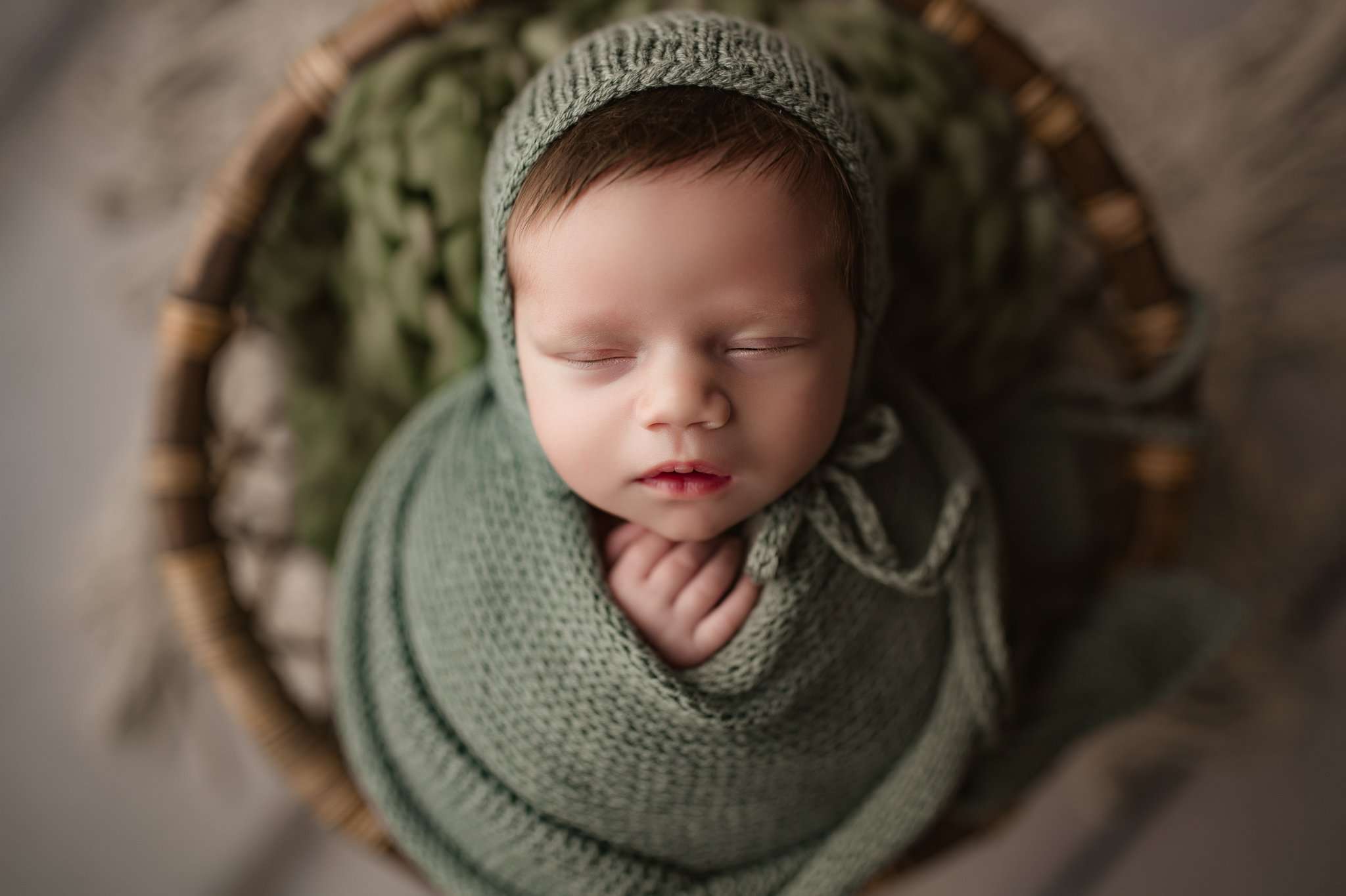 baby wrapped in a potato looking wrap with a green bonnet on while sleeping in a circular basket looking up at camera